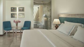 Quarto de hotel com uma cama grande, um espaço de estar com duas cadeiras, uma mesa pequena com um vaso de flores e um banheiro visível através de uma porta aberta.