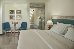 Quarto de hotel com uma cama grande, um espaço de estar com duas cadeiras, uma mesa pequena com um vaso de flores e um banheiro visível através de uma porta aberta.