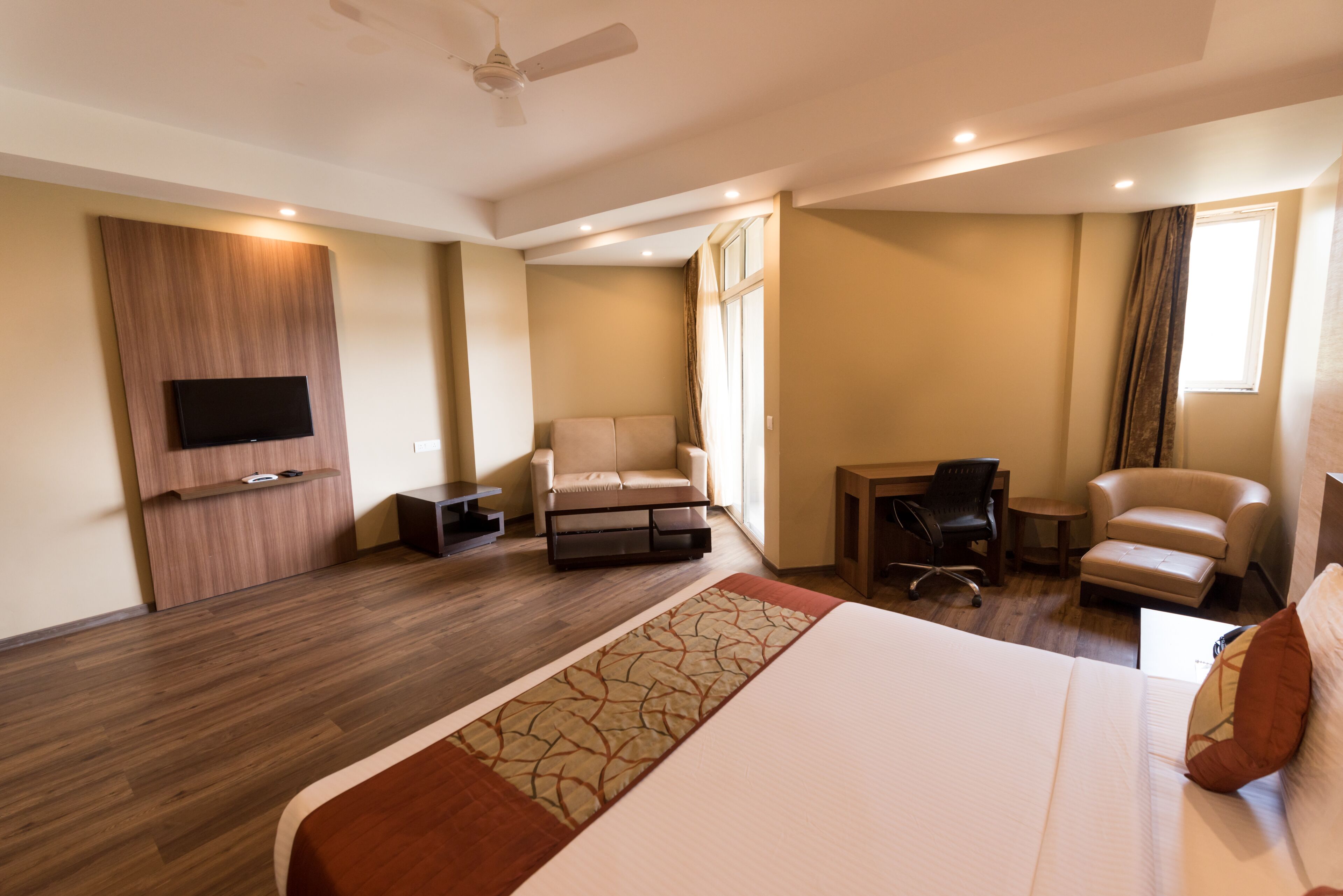 A modern hotel room with a large bed, a flat-screen TV mounted on the wall, a seating area with a sofa and chairs, and a small desk.