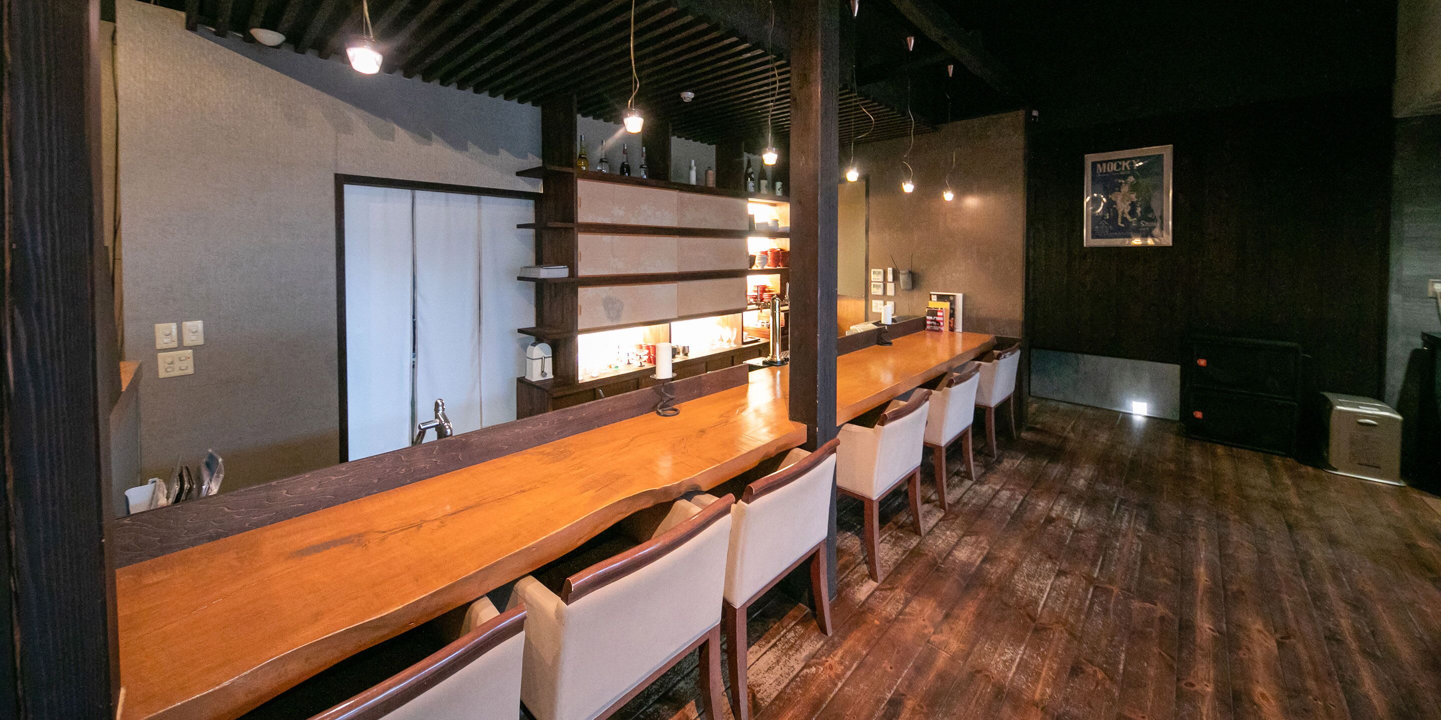 A bar area with wooden bar counter and white chairs, shelves with bottles, and a framed picture on the wall.