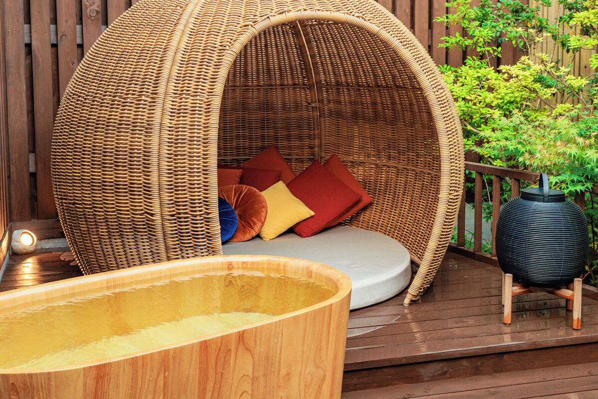 Une terrasse en bois avec un jacuzzi, un fauteuil suspendu en osier doté de coussins et une plante en pot.