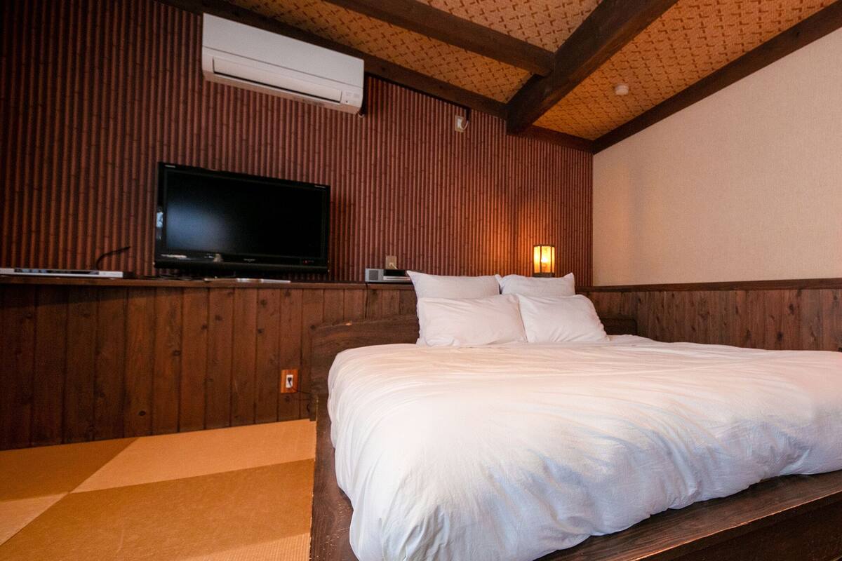 Une chambre à coucher avec une tête de lit en bois, un téléviseur à écran plat fixé au mur, une table de chevet avec une lampe et un grand lit avec une literie blanche.
