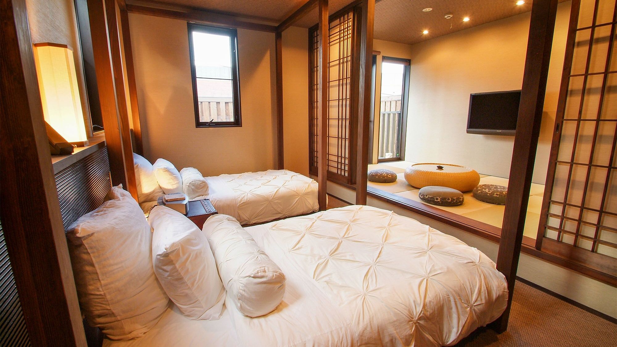 A hotel room with two beds, a bedside table, a lamp, a TV, and a view of the outside.