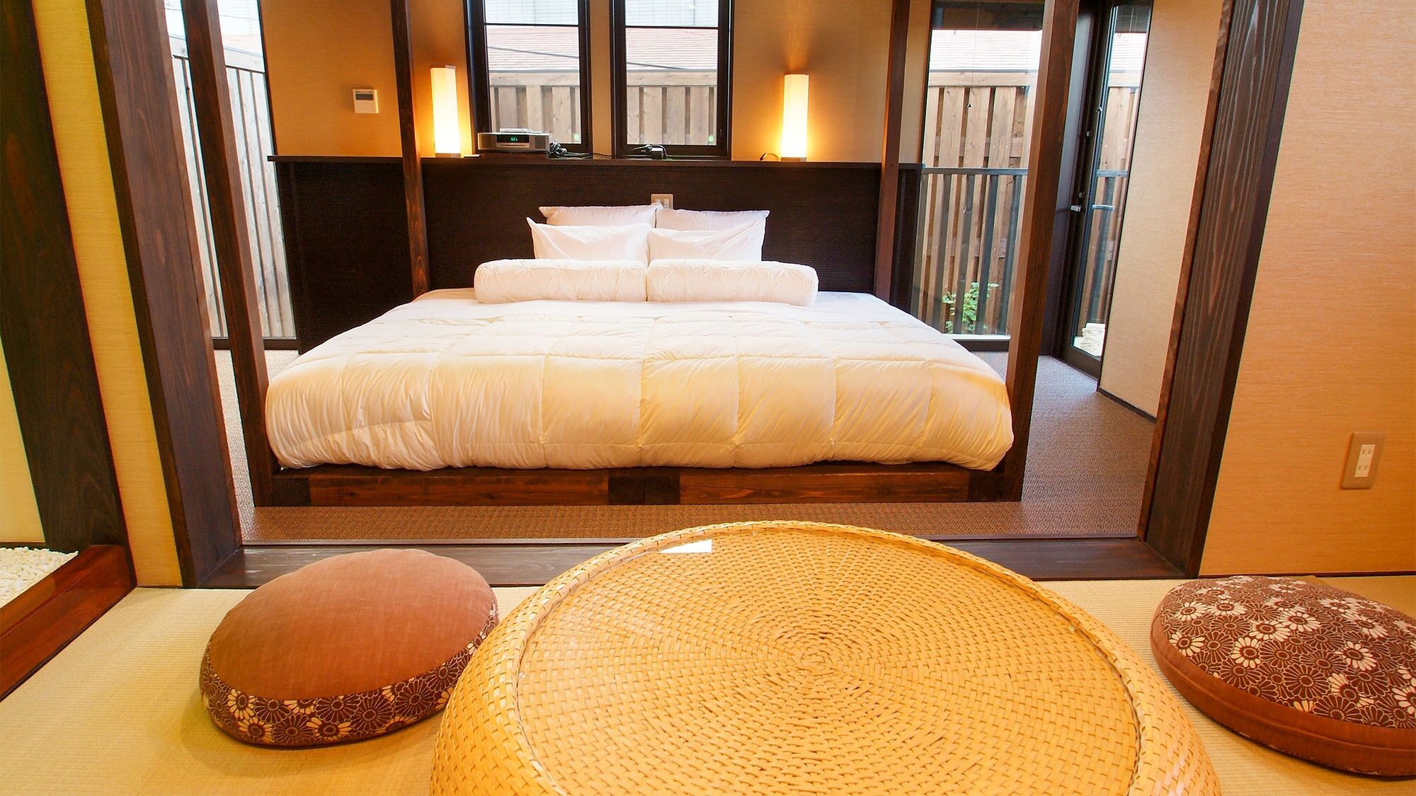 A hotel room with a large bed, a bedside table, a lamp, and a view of the outside.