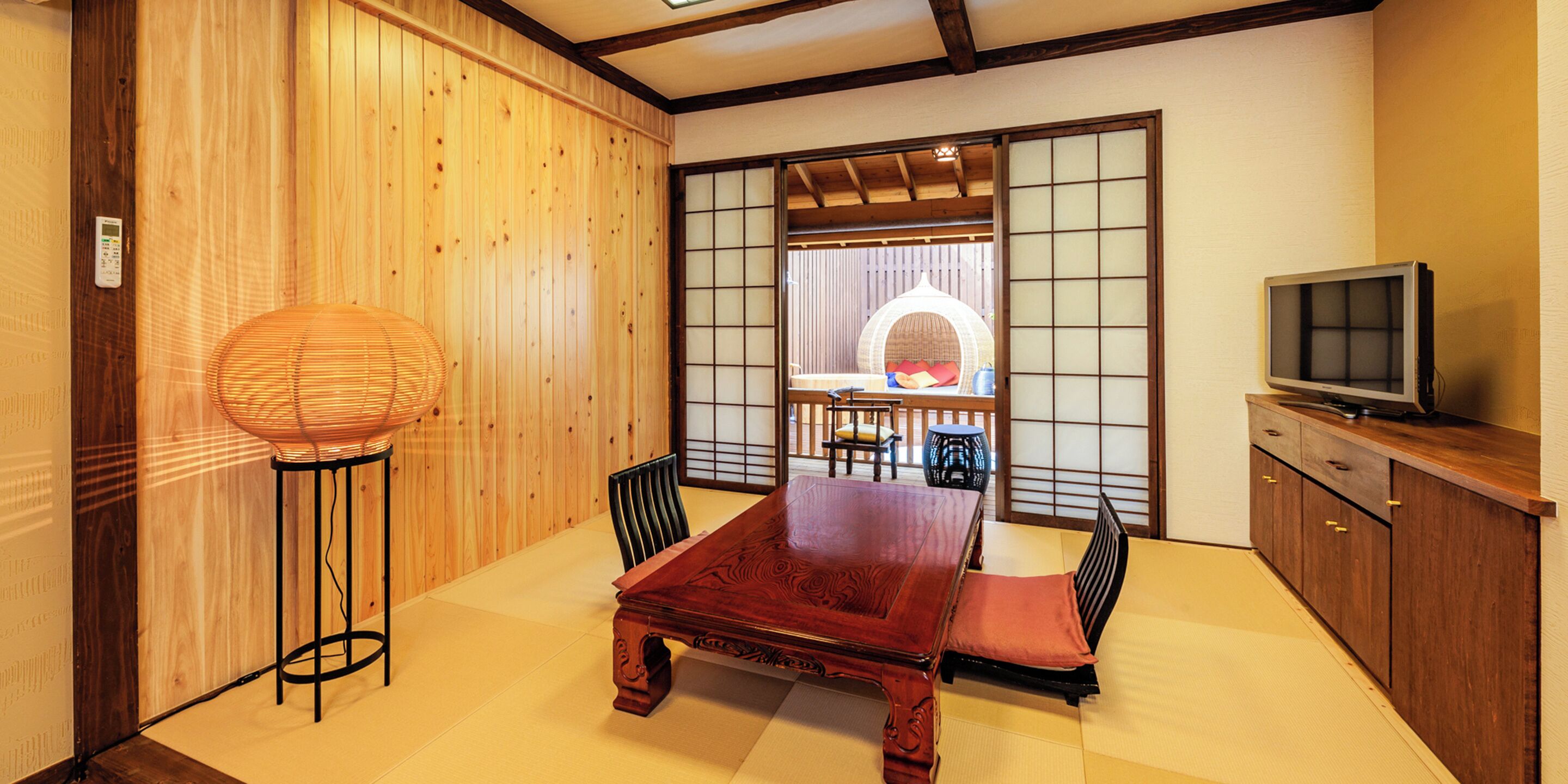 Una habitación tradicional japonesa con suelo de tatami, una mesa de madera baja, un banco de estera de tatami, una lámpara de pie tejida, un televisor sobre un mueble de madera y una puerta corredera que da a un balcón con vistas.