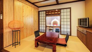 Una habitación tradicional japonesa con suelo de tatami, una mesa de madera baja, un banco de estera de tatami, una lámpara de pie tejida, un televisor sobre un mueble de madera y una puerta corredera que da a un balcón con vistas.