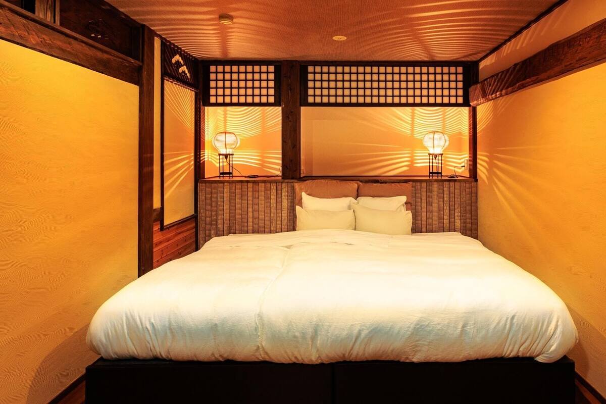 Une pièce de style japonais traditionnel, avec un plafond en bois, un lit recouvert de draps blancs et deux lampes fixées au mur.