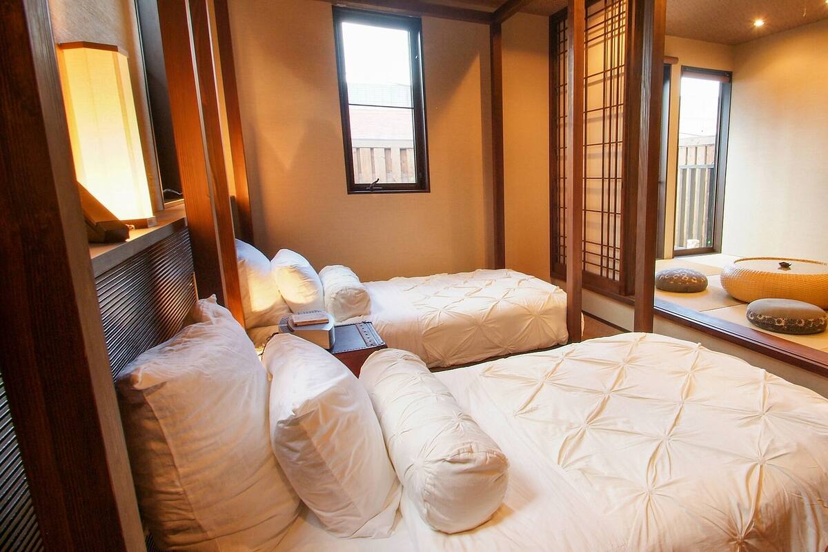Une pièce de style japonais traditionnel, avec un sol recouvert de tatamis, deux nattes de tatami, un lit bas en bois, un paravent coulissant de type shoji, une petite table en tatami avec un bol et un coussin, et un téléviseur fixé au mur.