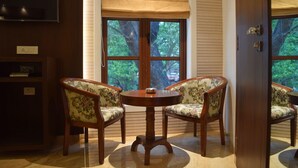A room with a round wooden table and two floral-patterned chairs, a large window with a beige blind, and a television mounted on the wall. - South Coast Hotels Thane (Thane)