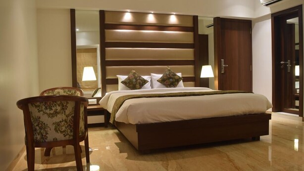 A hotel room with a large bed, a nightstand, two chairs, a mirror, and a wardrobe. - South Coast Hotels Thane (Thane)