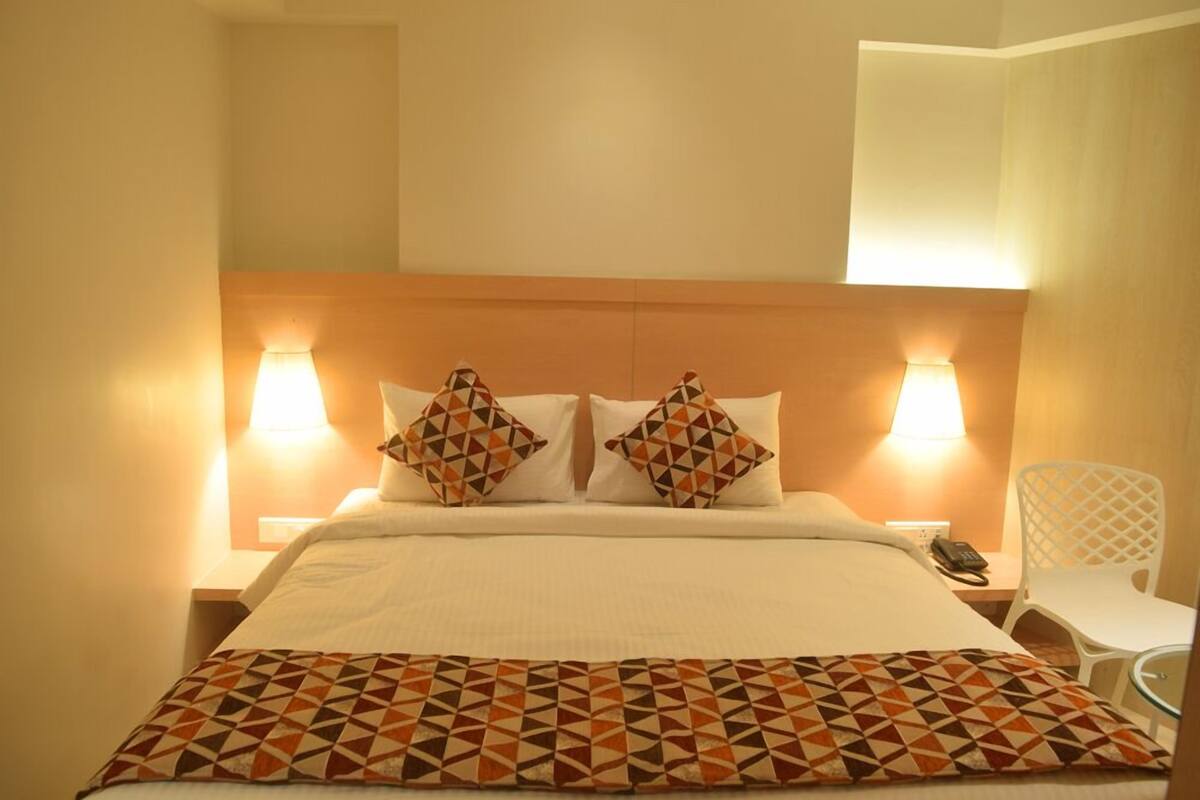 A hotel room with a large bed, two patterned pillows, bedside lamps, a white chair, and a phone on the nightstand.