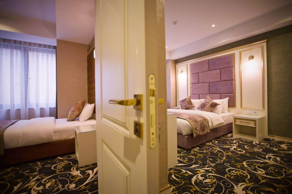 Ein Hotelzimmer mit zwei Betten, einem großen Fenster mit transparenten Vorhängen, einem gemusterten Teppich und einer Tür mit goldener Griff.