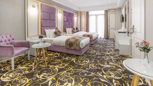 Una habitación de hotel con una cama grande, una mesita con un florero, un sillón morado y un espejo.