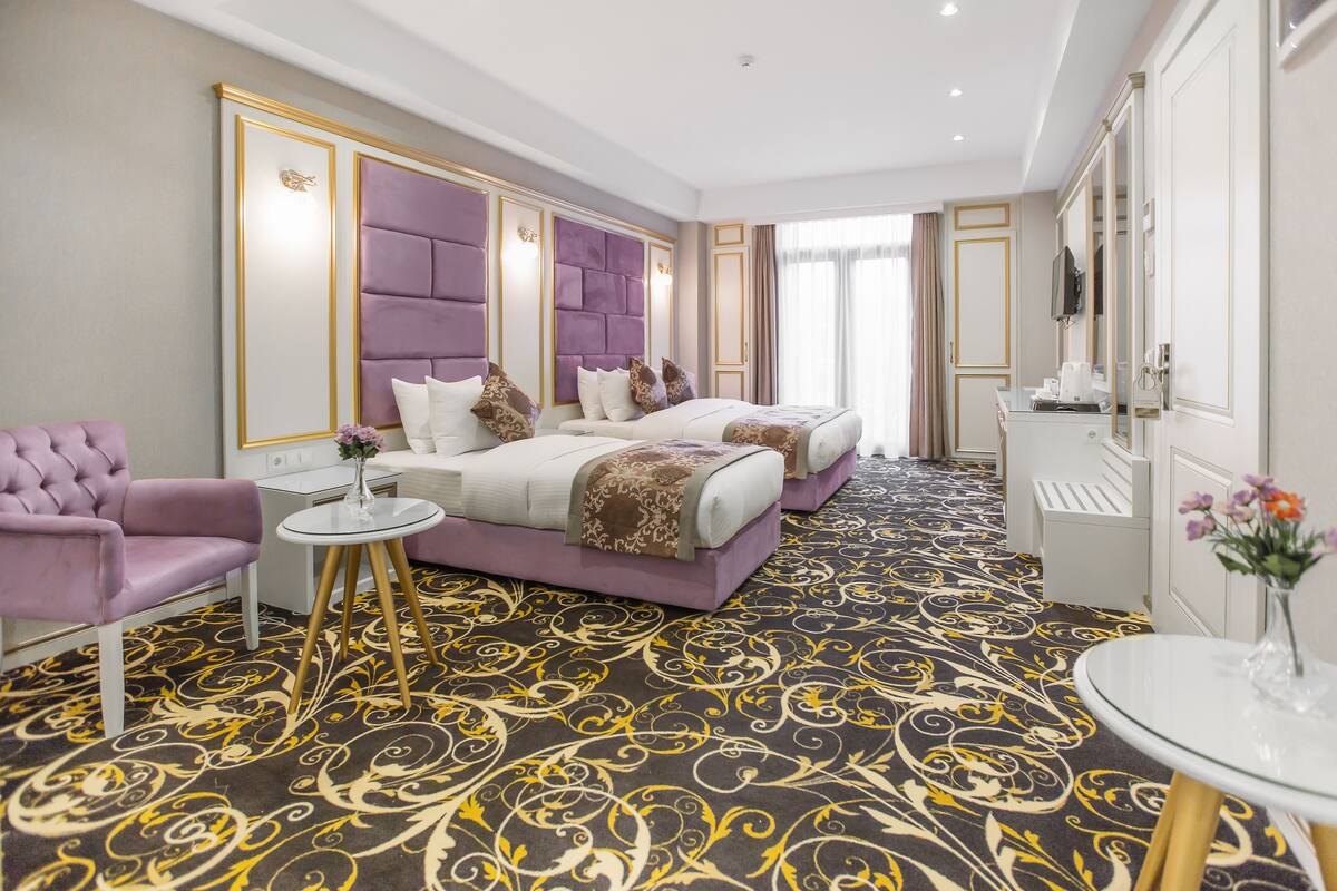 Ein Hotelzimmer mit einem großen Bett, einem kleinen Tisch mit einer Vase, einem violetten Sessel und einem Spiegel.