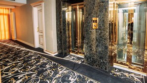 Un hall de hotel con piso de mármol, un ascensor de vidrio y una puerta con picaporte dorado.