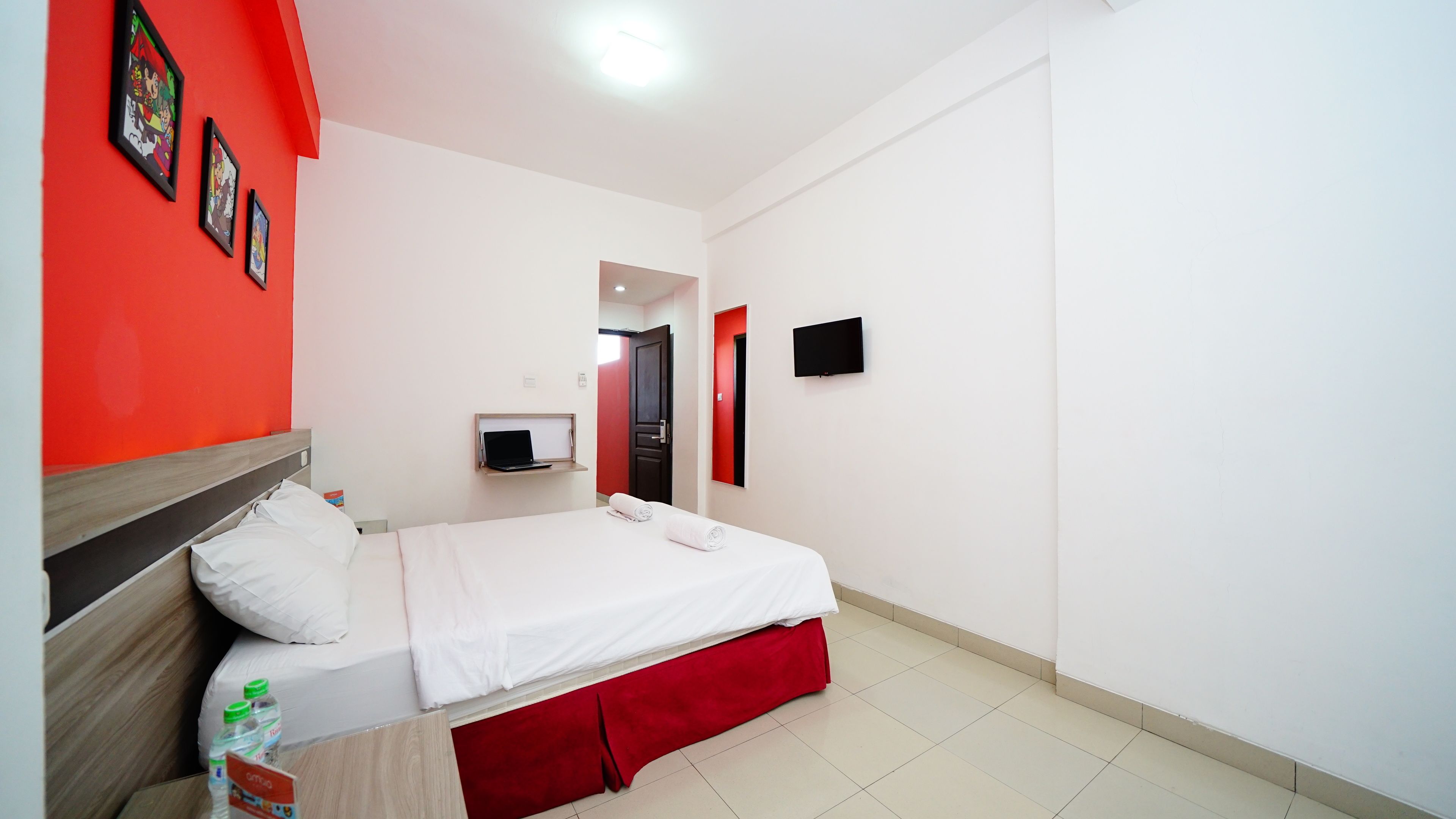 Habitación de hotel con cama, escritorio con laptop, televisor en la pared y una pared de acento roja.