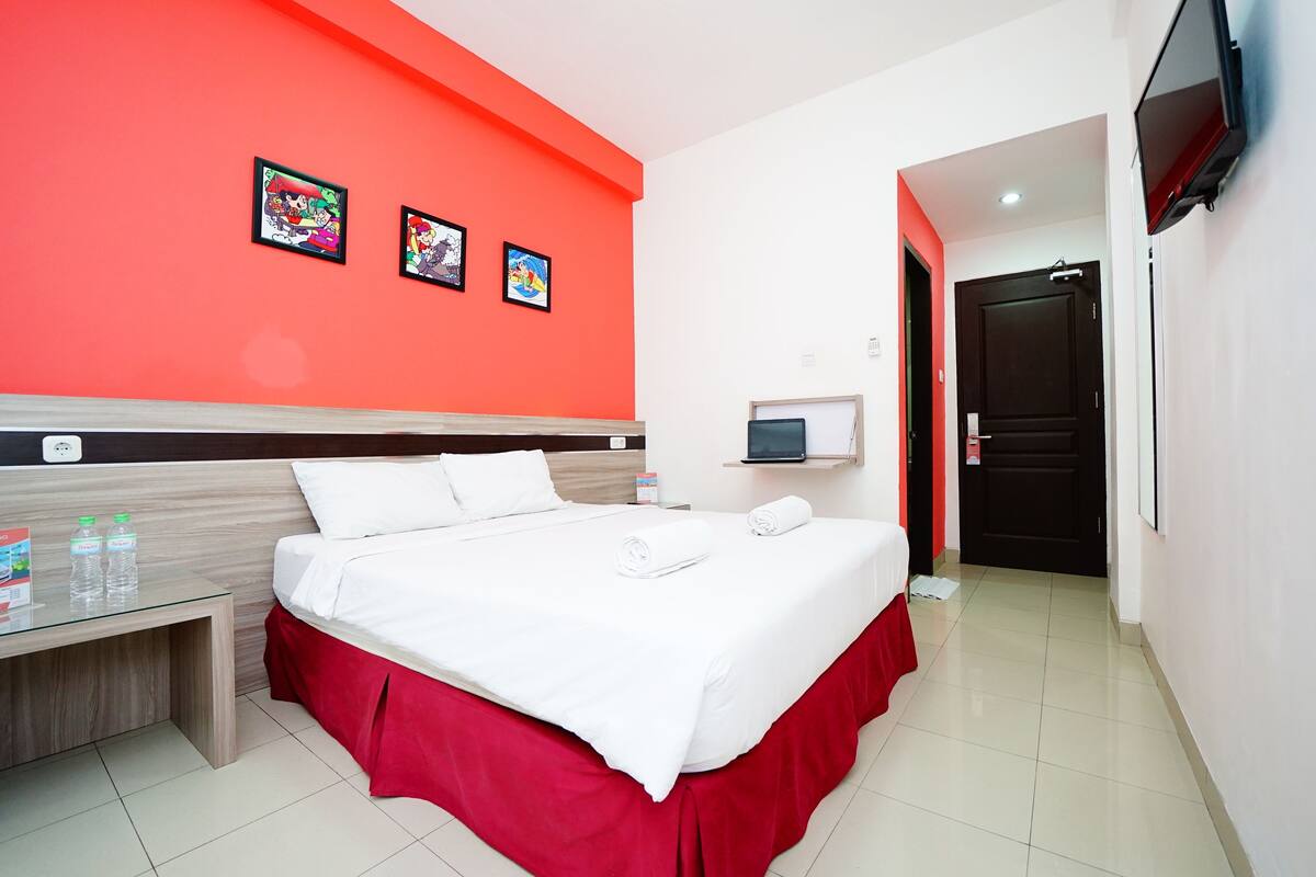 A hotel room with a bed, a bedside table with water bottles, a TV, and three framed pictures on the wall.