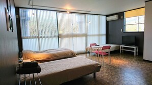 A bedroom with a bed, a small table with a chair, a TV, and large windows with blinds.