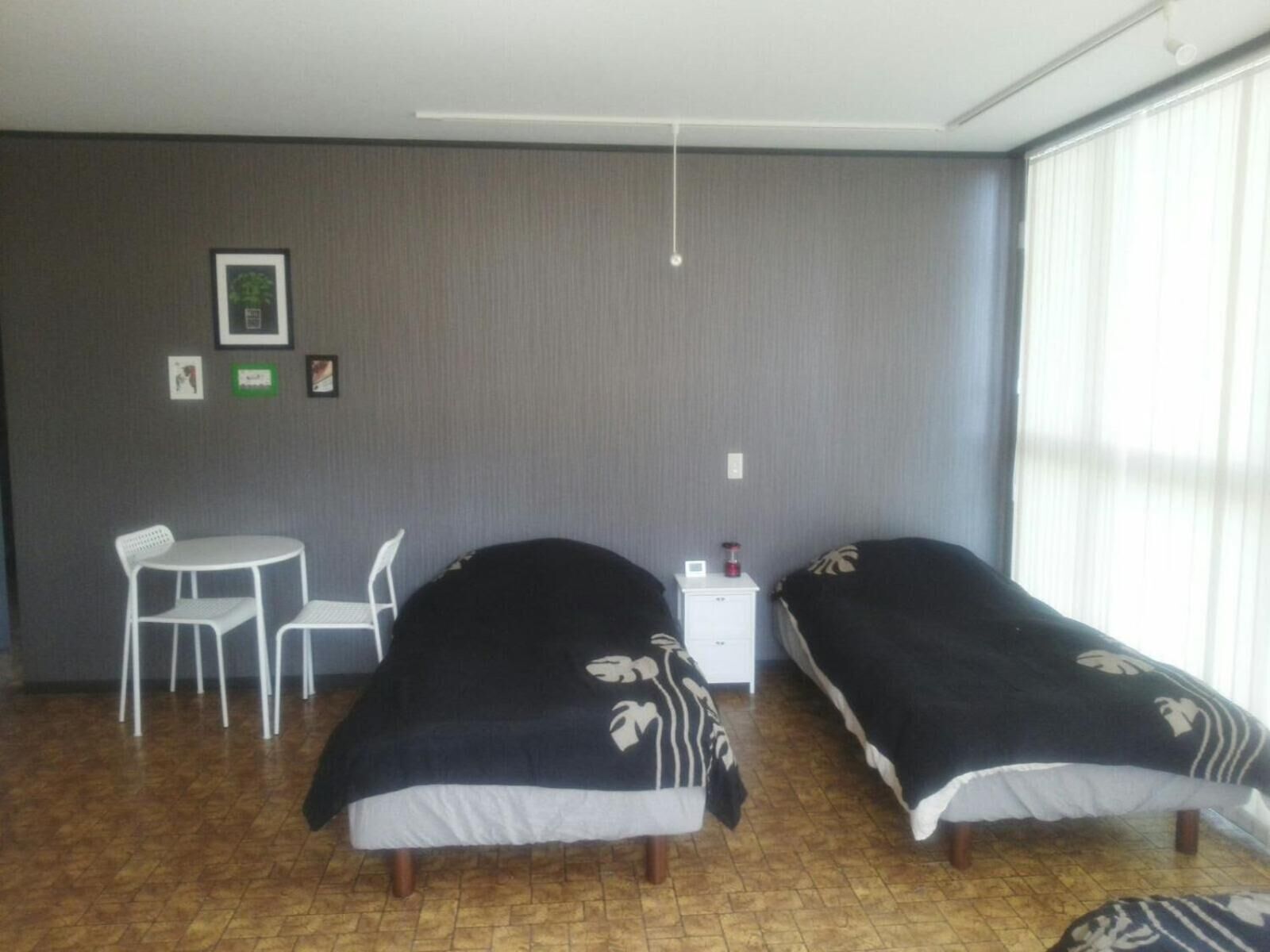 A room with two single beds, a small table with a chair, a bedside table, and a window with blinds.