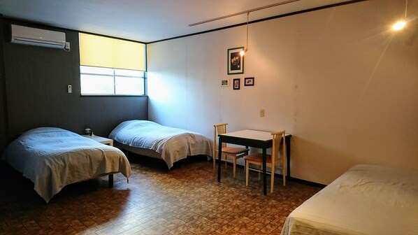A small, compact room with two single beds, a small table with a chair, and a window providing natural light.