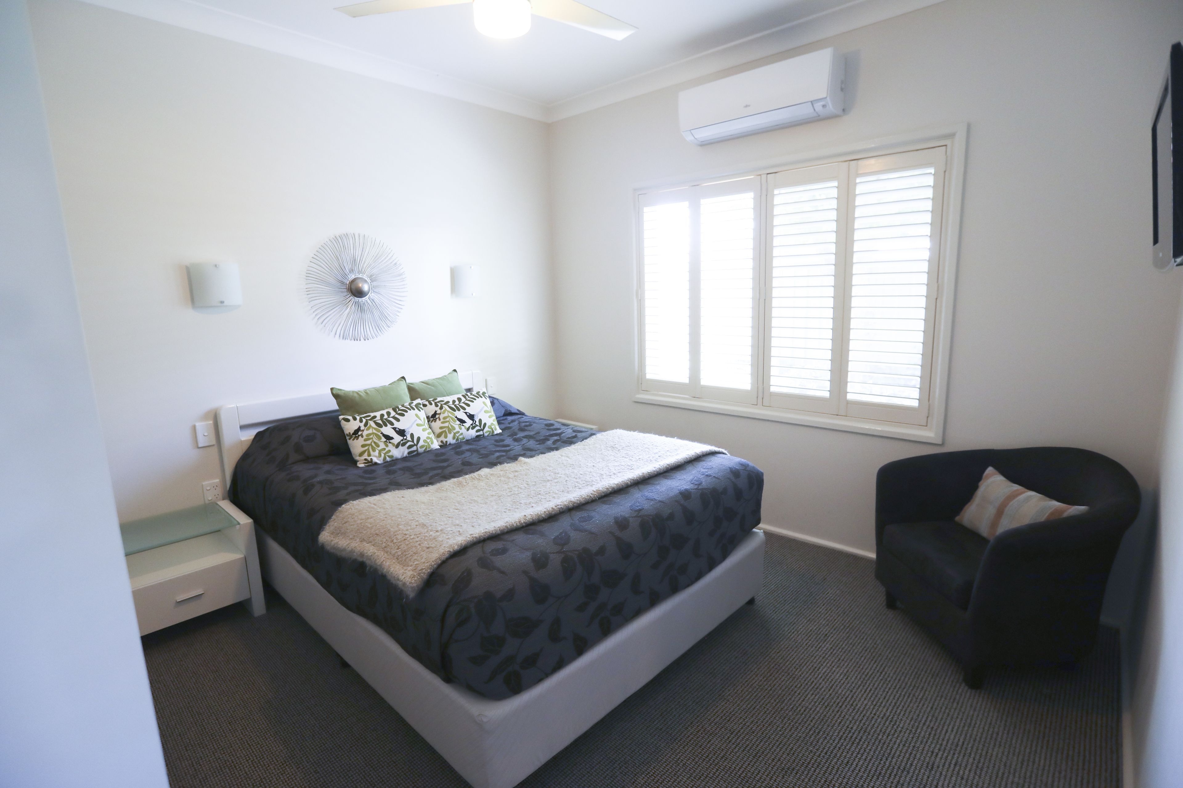 A neatly arranged bedroom with a bed, bedside table, chair, and a window with blinds.