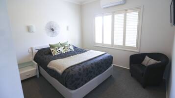 A neatly arranged bedroom with a bed, bedside table, chair, and a window with blinds.