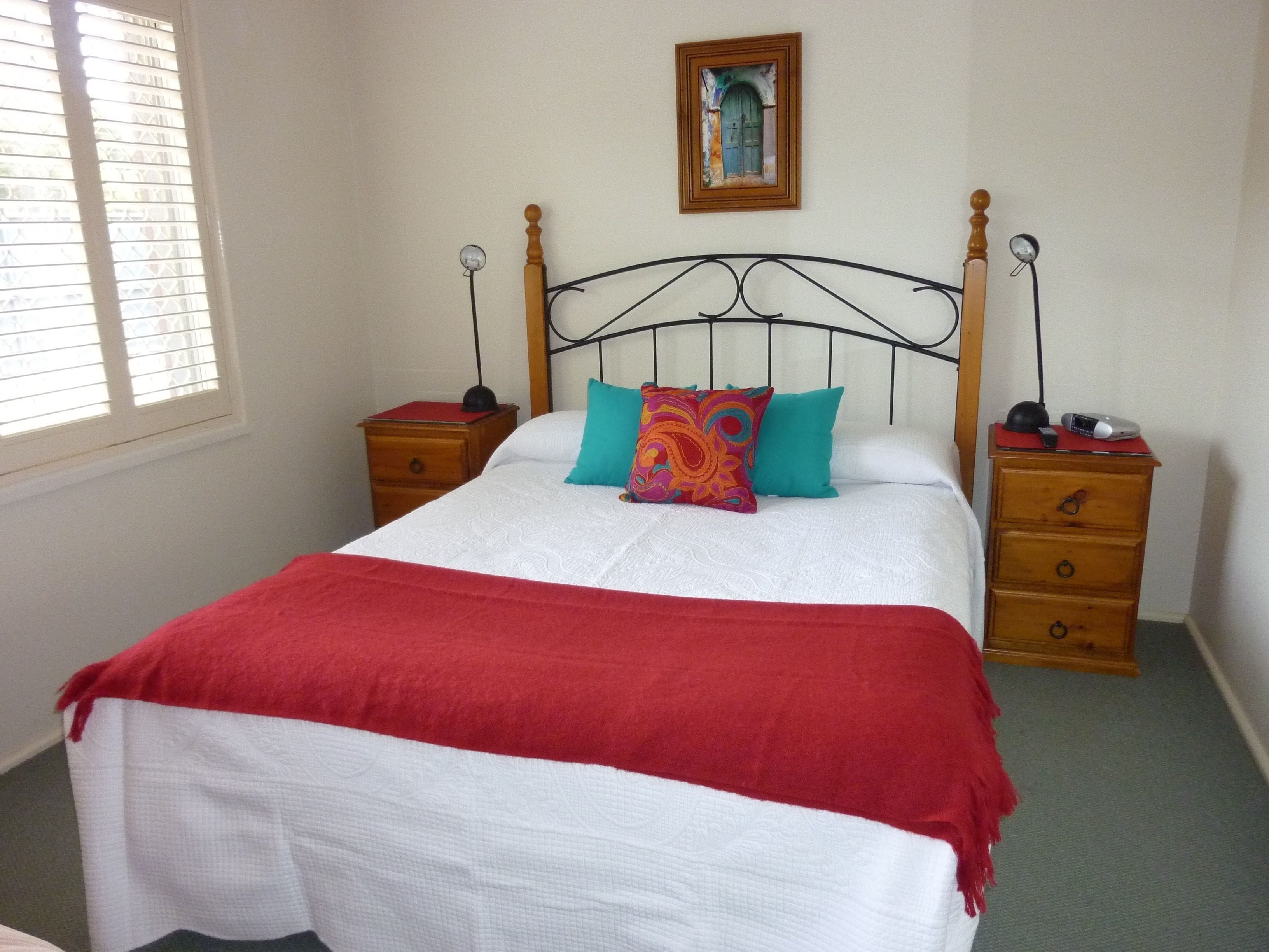 A neatly made bed with a red blanket, a wooden headboard, and a colorful pillow. There are two bedside tables with lamps and a small drawer. A framed picture hangs on the wall above the bed.