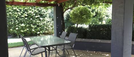 Een buitenpatio met een glazen tafel en metalen stoelen, omgeven door groen en een pergola.
