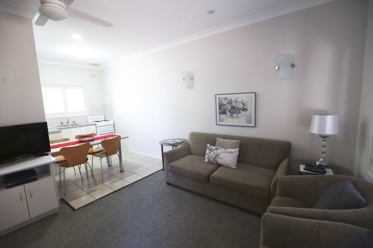 Ein Wohnzimmer mit Sofa, Fernseher, Esstisch mit Stühlen, Küchenzeile und Deckenventilator.