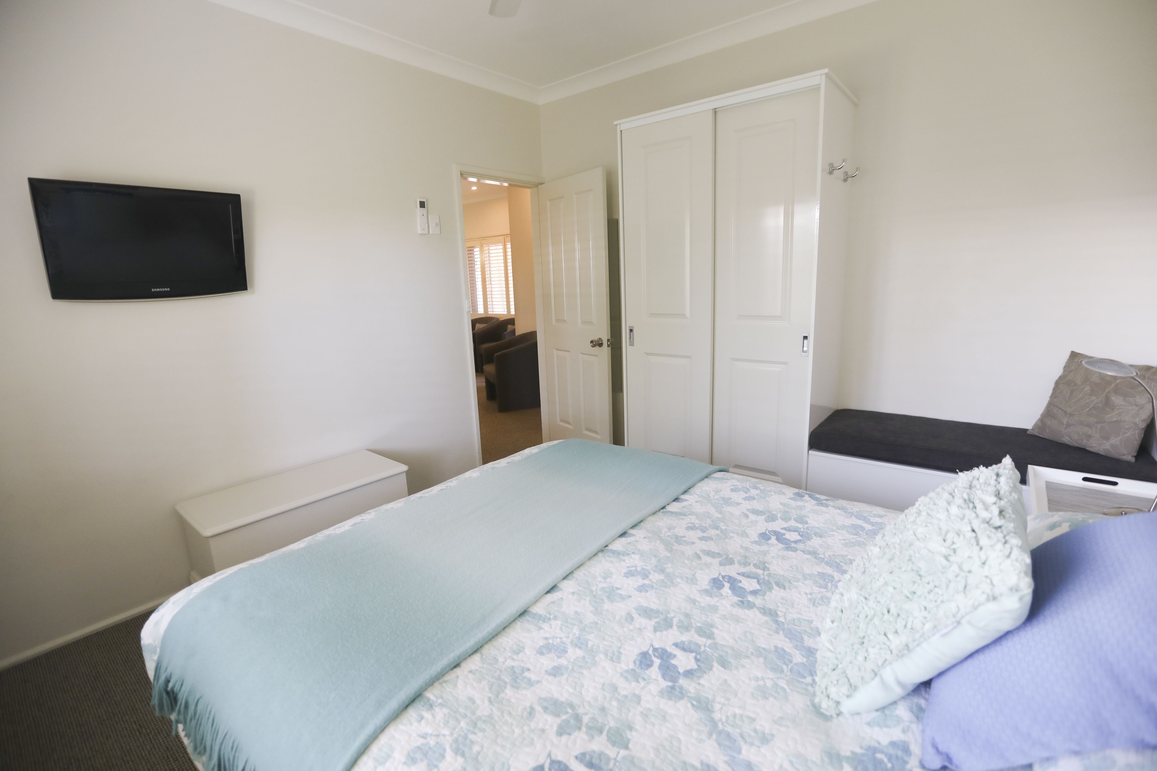 A bedroom with a bed, a TV mounted on the wall, a wardrobe, and a bench with cushions.