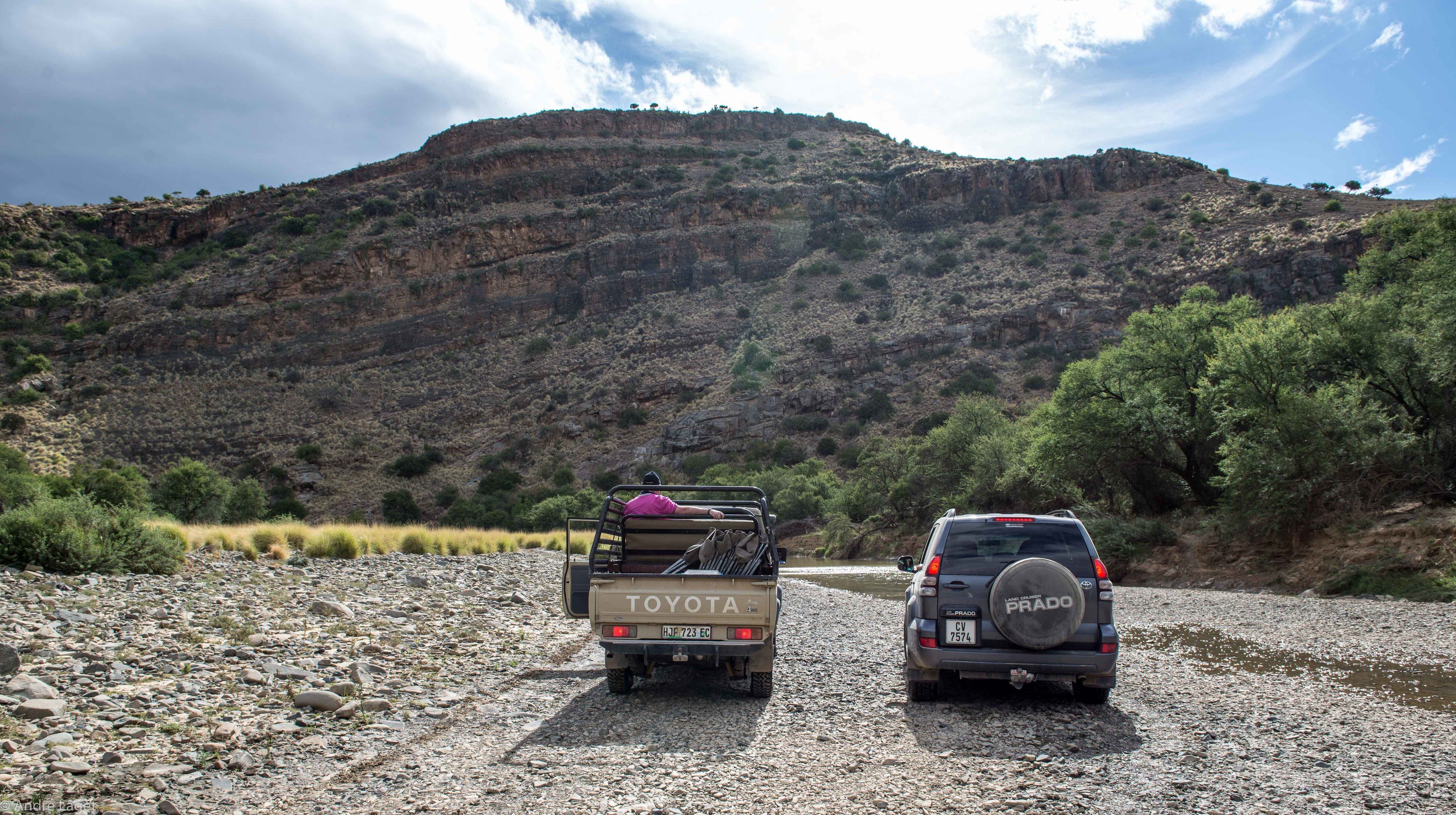 Dos vehículos estacionados en un camino de grava con montañas al fondo.