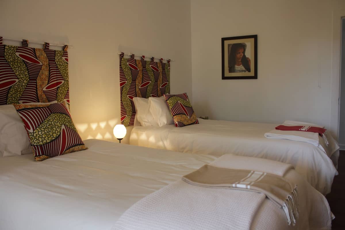 Ein Hotelzimmer mit zwei Betten, jedes mit gemusterten Kissen und einer weißen Decke. An der Wand hängt ein gerahmtes Bild und auf der linken Seite steht eine Lampe.