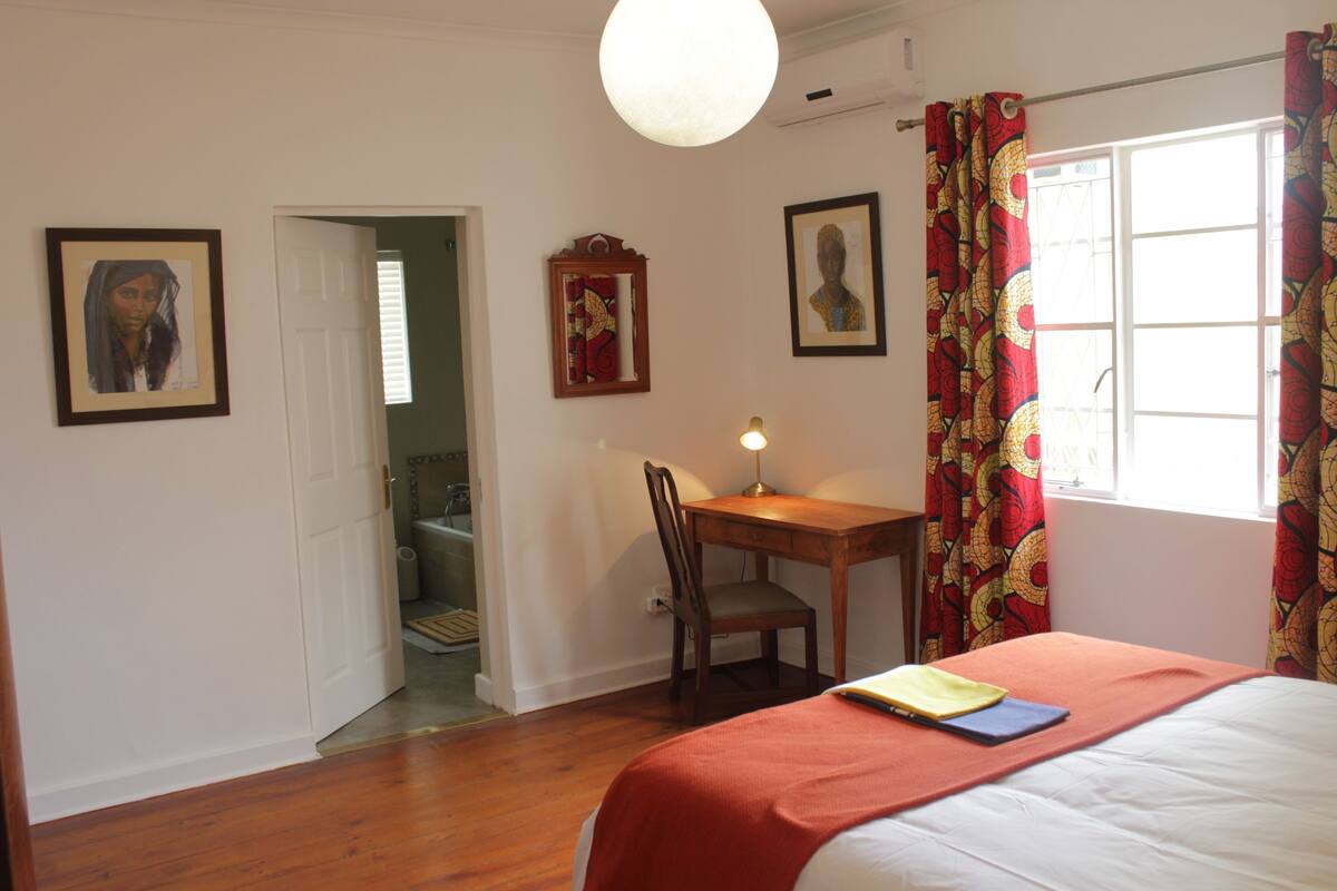 Ein Schlafzimmer mit Bett, Schreibtisch und Stuhl. An der Wand hängen gerahmte Bilder.