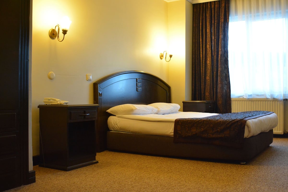 A hotel room with a large bed, a bedside table, a lamp, and a window with curtains.