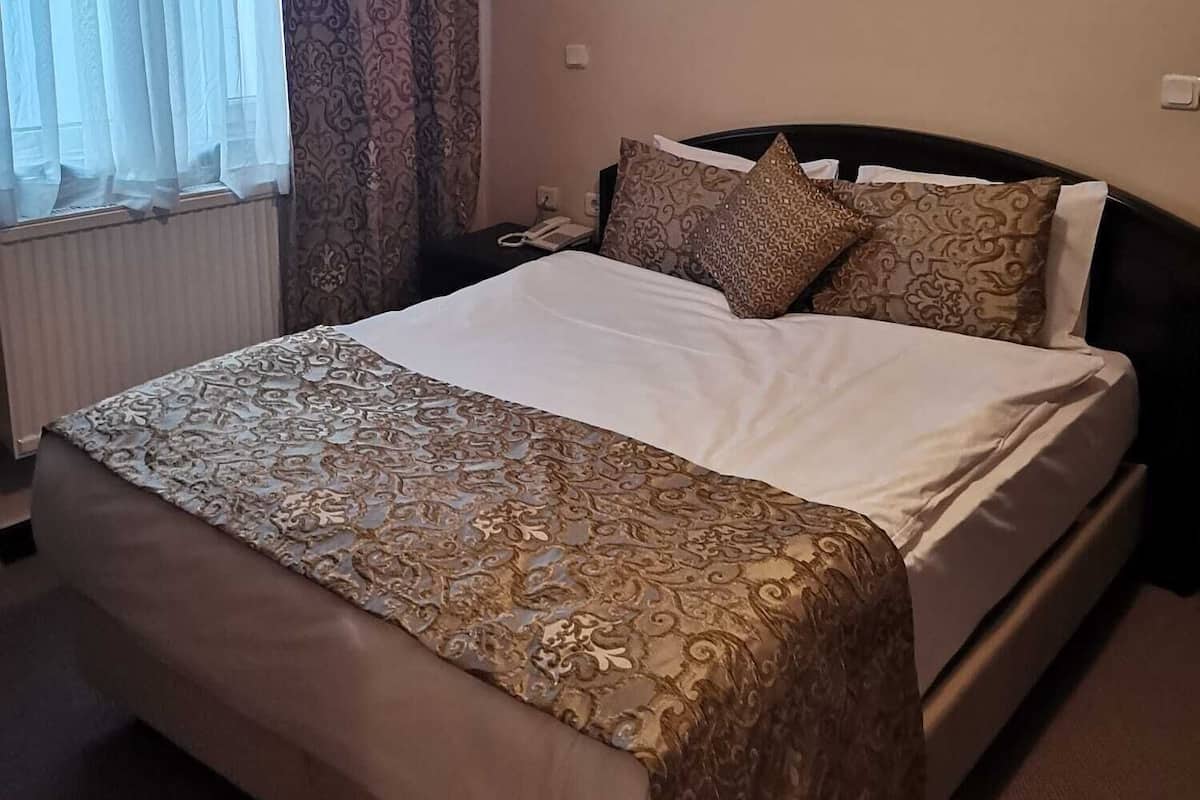 A hotel room with a large bed, a bedside table, a nightstand, a window with sheer curtains, and a wall-mounted light fixture.