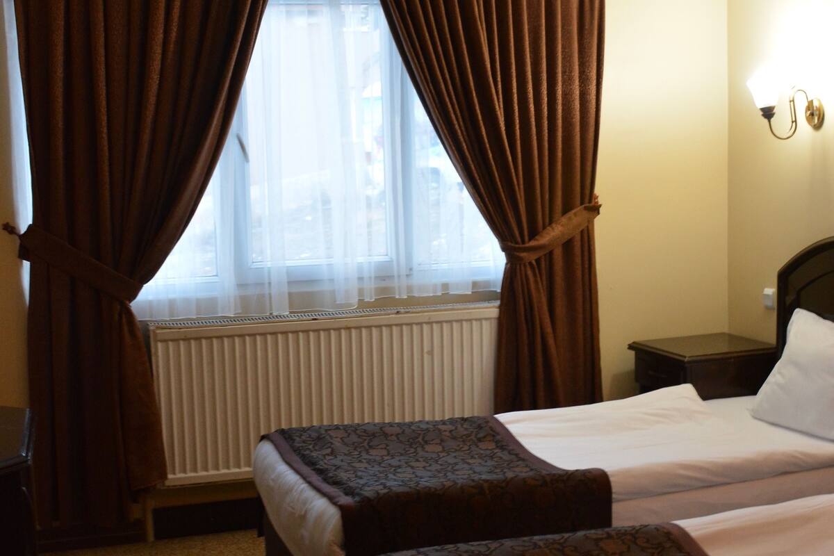 A hotel room with two beds, a radiator, a window with curtains, and a bedside table.