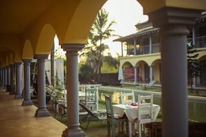 A covered outdoor area with columns, tables, and chairs, overlooking a pool and a building with arches.