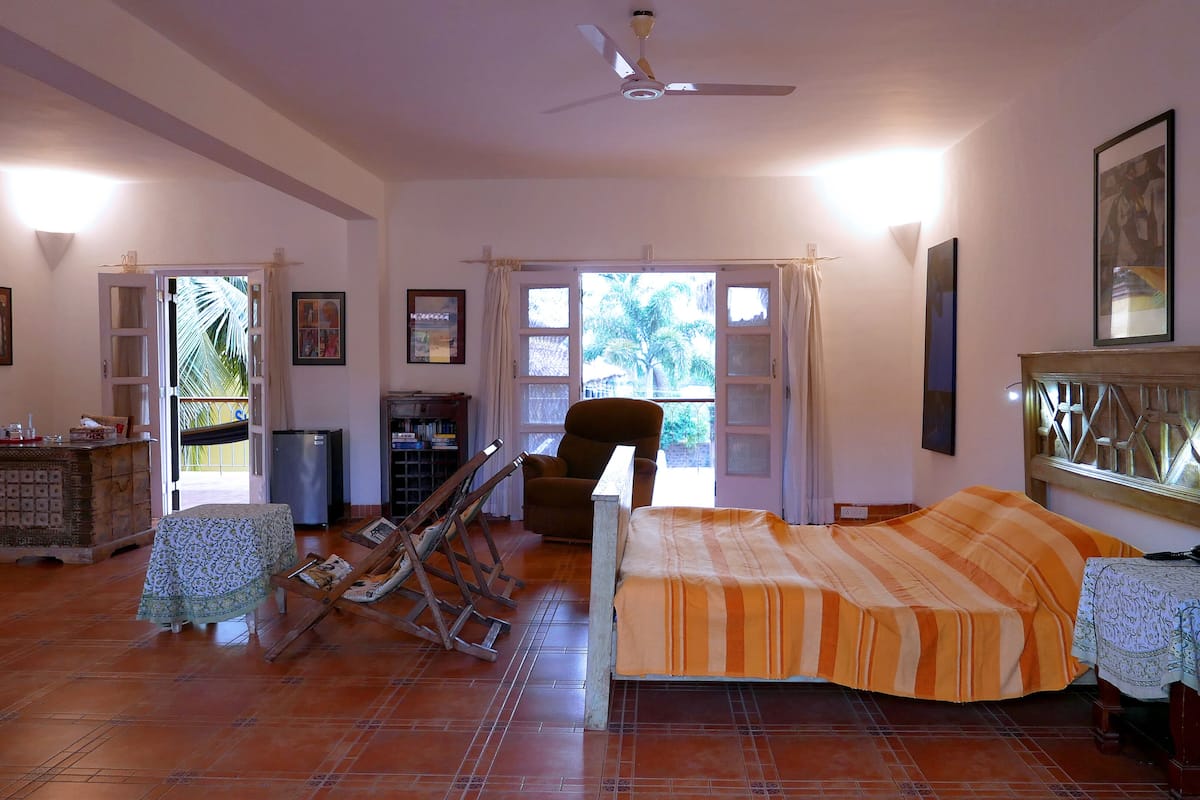 A spacious living room with a sofa, a chair, a small table, and a television. There is a ceiling fan and a window with curtains.