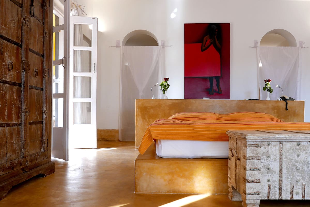 A room with a wooden door, a bed with an orange striped blanket, a white bedside table, a red artwork on the wall, and a white vase with a flower.