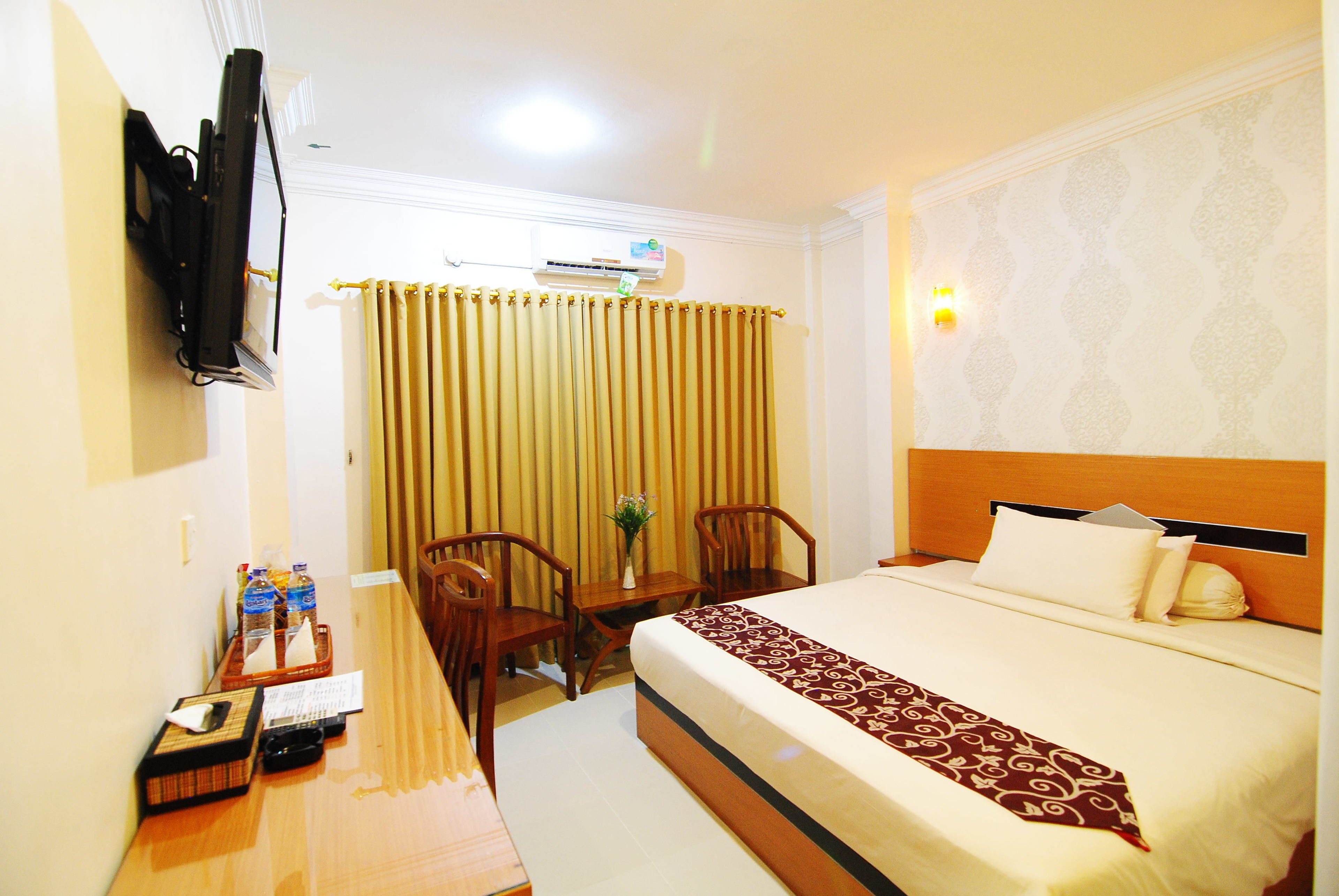 Quarto de hotel com cama, escrivaninha com cadeira, televisão e janela com cortinas.