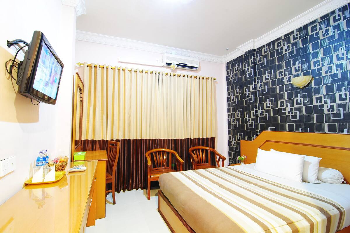 A hotel room with a bed, a desk with a television, two chairs, a window with curtains, and a patterned wall.