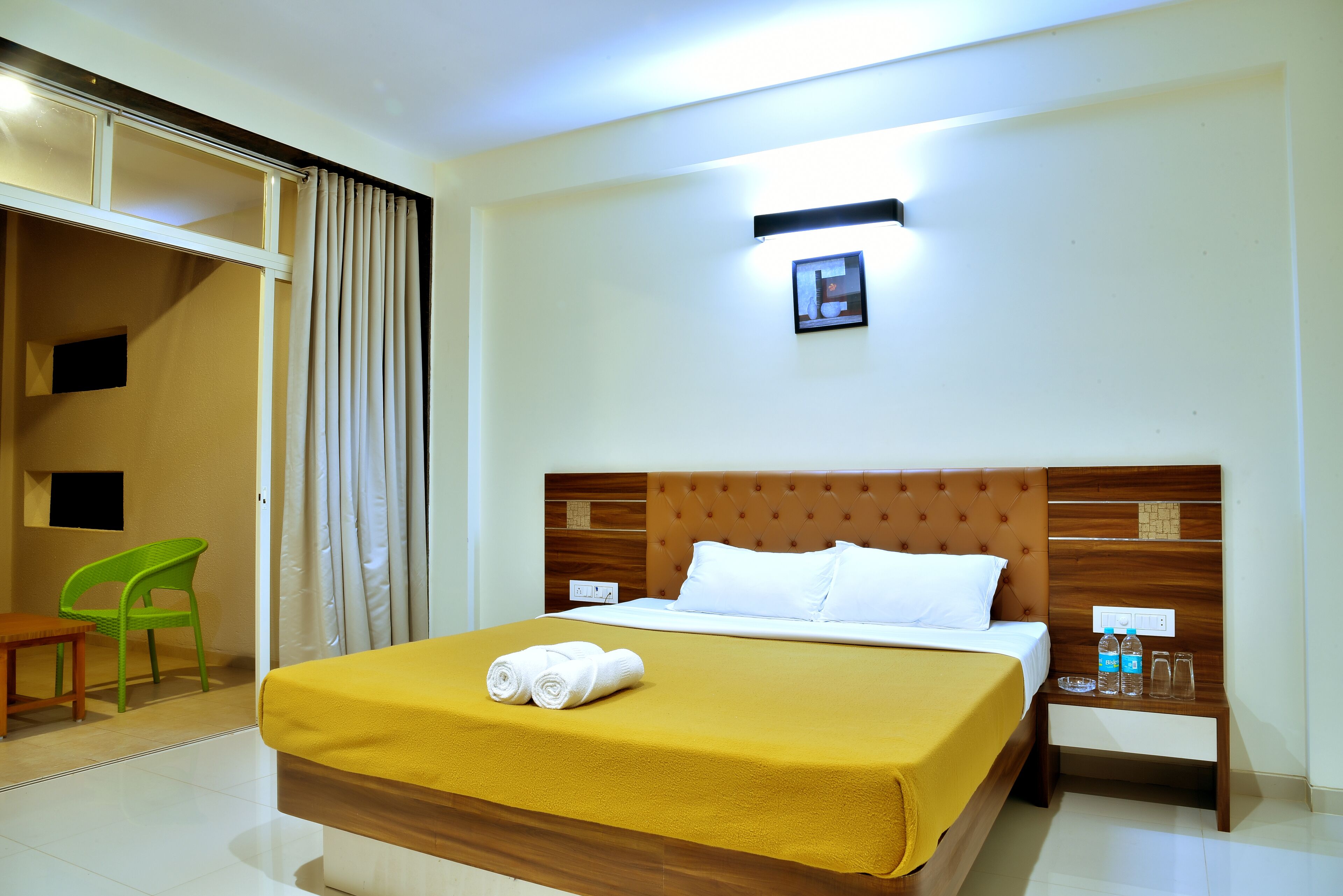 A hotel room with a bed, a nightstand, a green chair, and a sliding door.