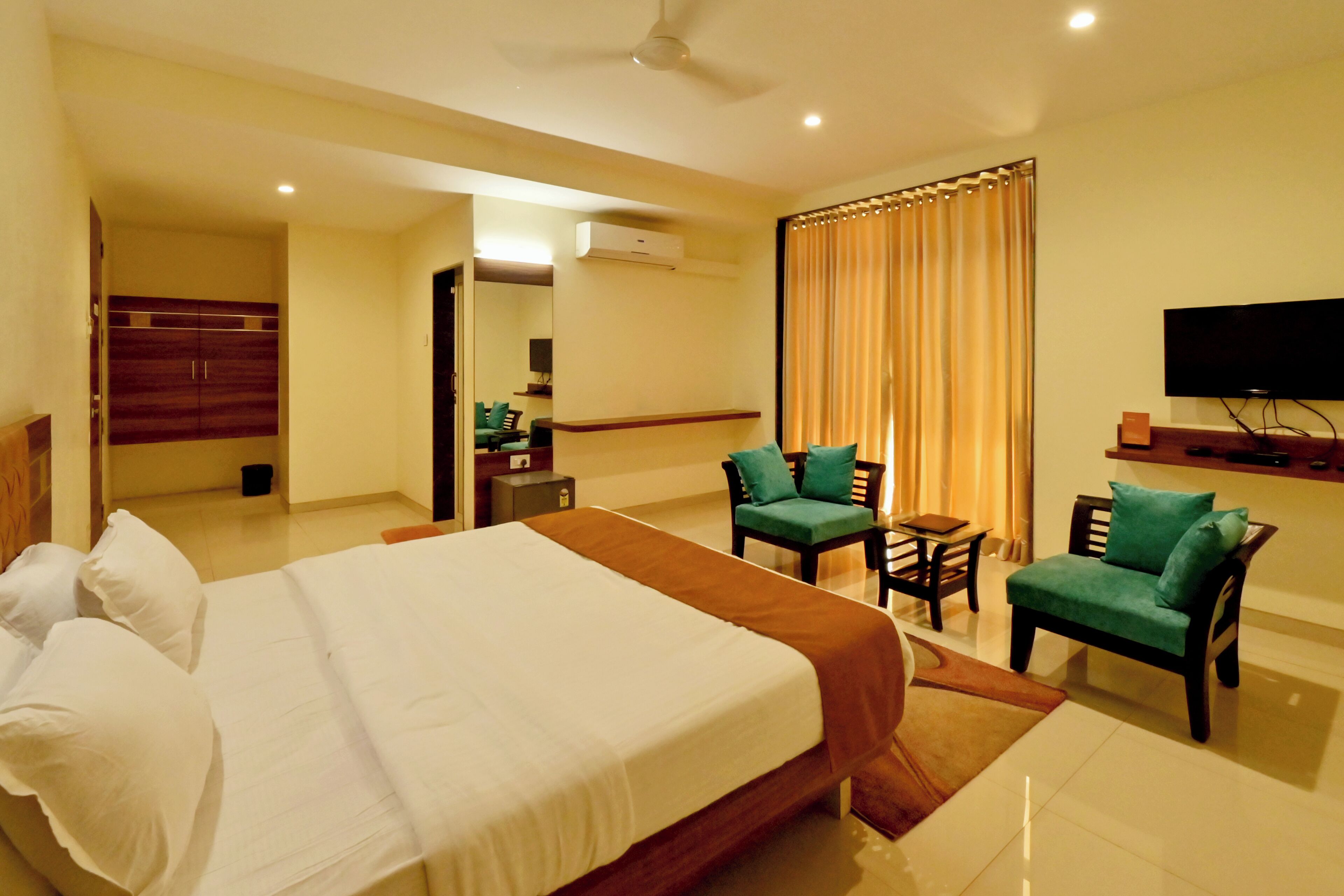 A hotel room with a large bed, two green armchairs, a small table, a TV, and a mirror.