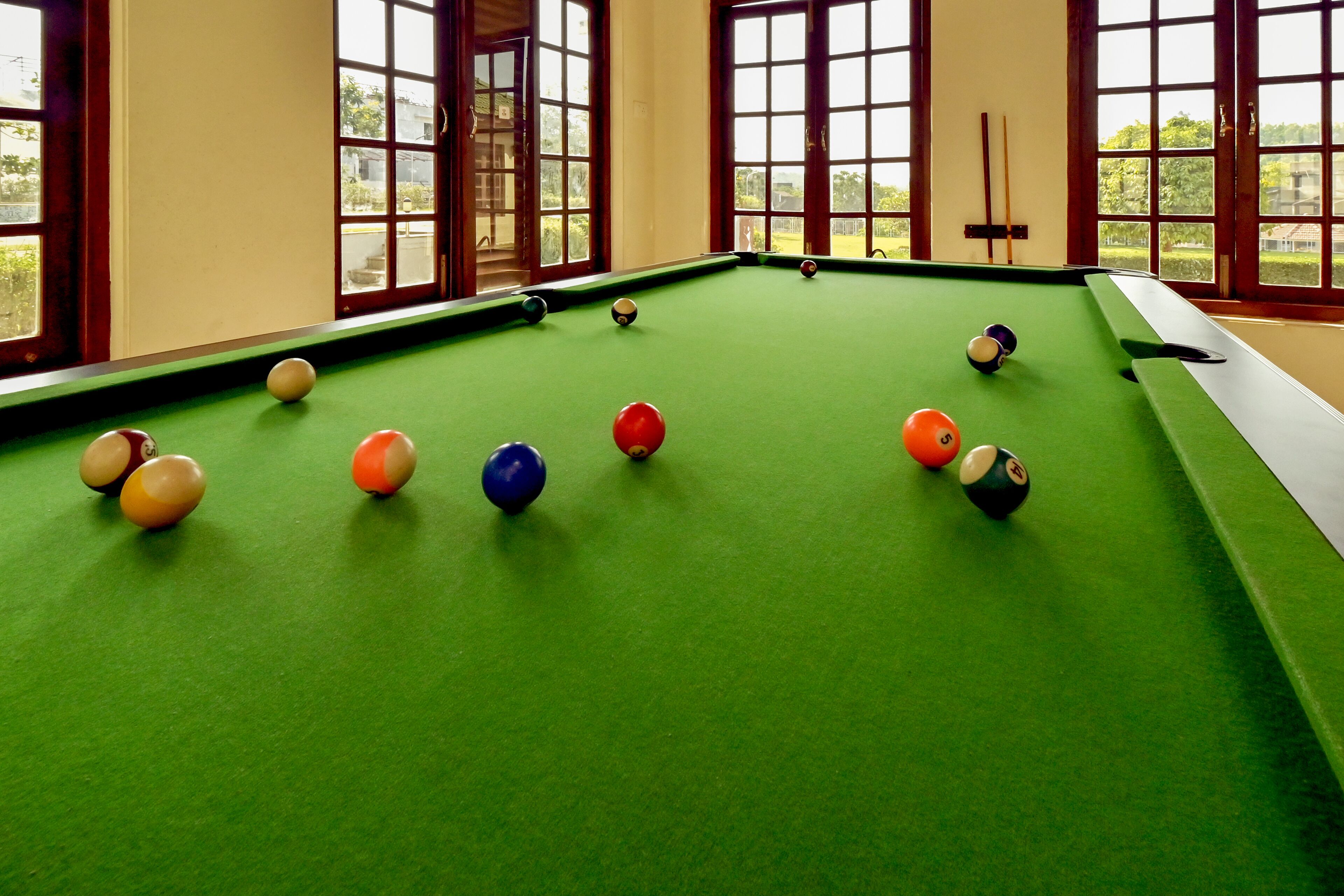 Una mesa de billar con bolas dispuestas en forma de triángulo, amplias ventanas que dan a vistas de vegetación, y un soporte para tacos de billar en la pared.