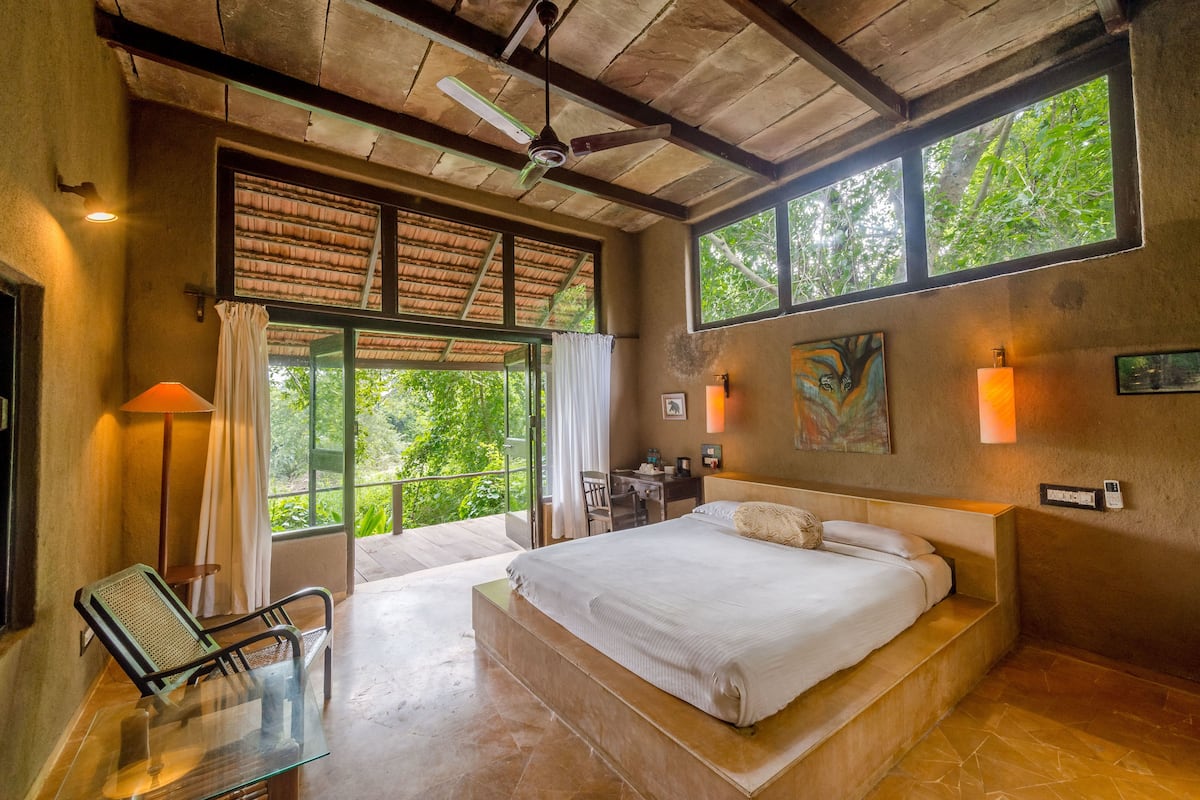 Un dormitorio amplio con una cama grande, una mesita de vidrio, un escritorio y una silla. Hay ventanas con cortinas, un ventilador de techo y obras de arte en las paredes.
