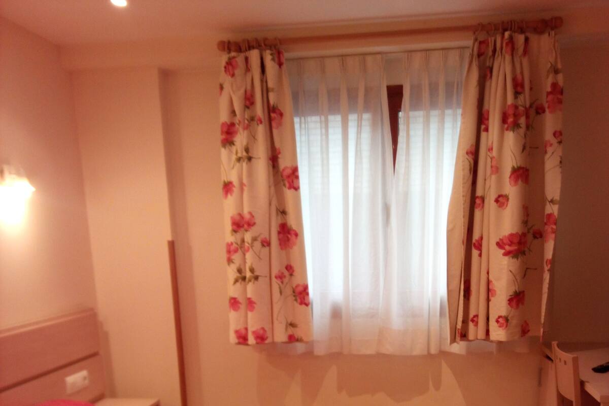 A bedroom with a bed, a nightstand, a desk, and a window with floral curtains.