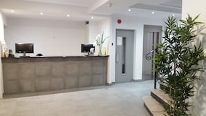 Zona de recepción con mostrador, dos monitores, una planta en maceta y una puerta que da a otra habitación.