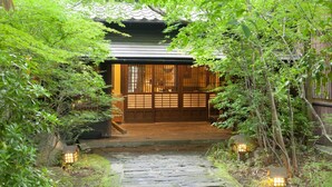 A traditional Japanese gate leading to a wooden building with a tiled roof, surrounded by lush greenery and stone pathways. - Yamashinobu (Minamioguni)