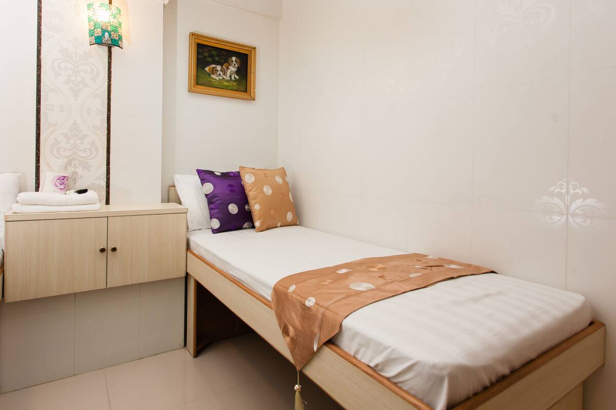Une petite chambre à coucher, bien rangée, avec un lit simple, une table de chevet en bois, une serviette et une lampe fixée au mur.