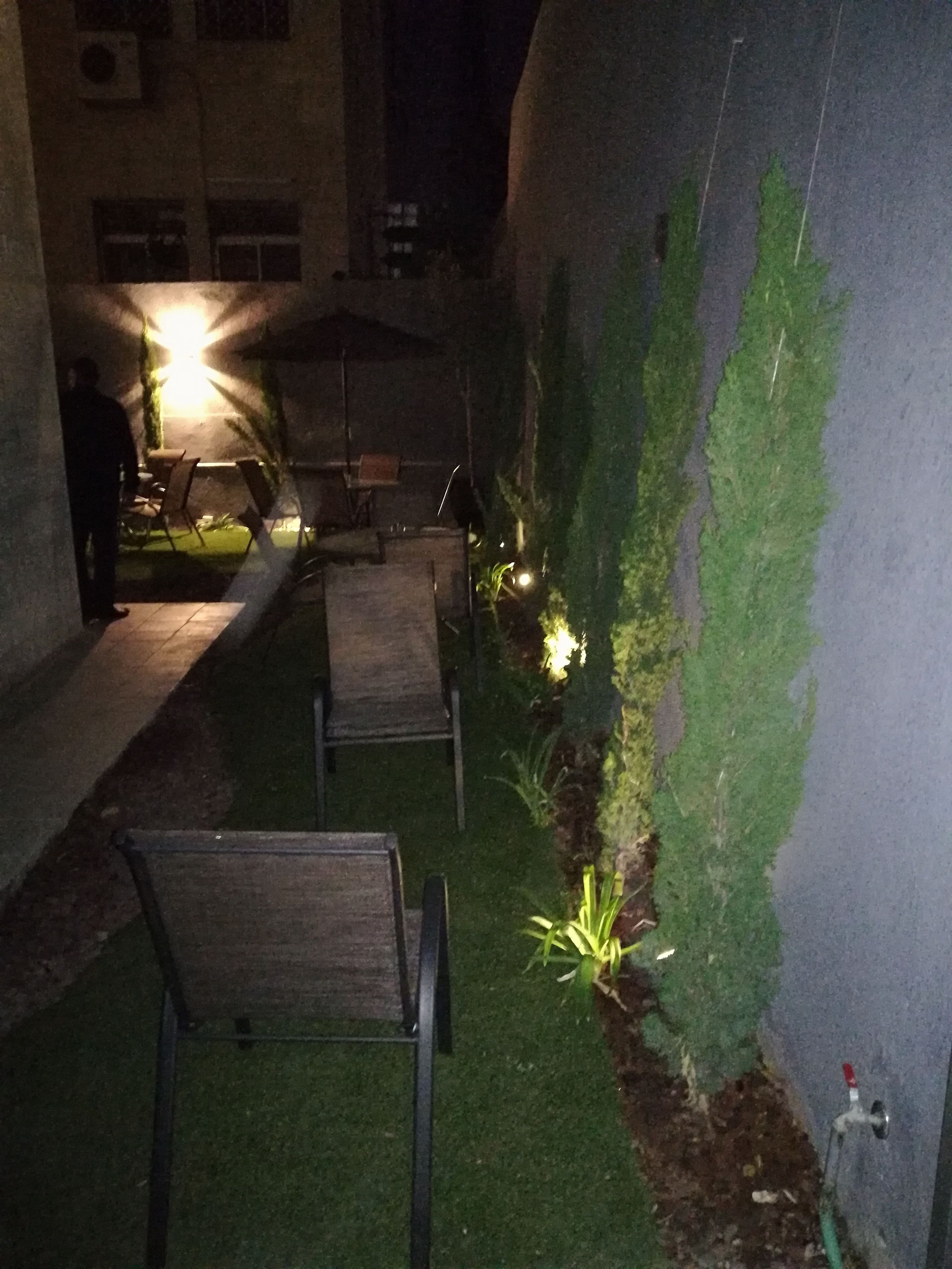 Een terras met stoelen en tafels, een kleine tuin met planten, en een gebouw op de achtergrond.